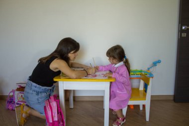 Genç bir anaokulu öğretmeninin pembe önlüklü küçük bir kıza not defterini boyamayı öğretmesi. Okula dönüş ve ilkokulda gerçekleştirilen faaliyetlere atıfta bulun