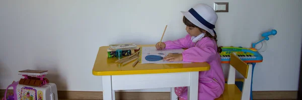 Adorable little girl with pink hat and apron playing with pencil colors in a notebook. Back to school and first day of kindergarten. Horizontal banner