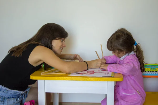 Image of a young mom teaching her daughter to color with pencils. Little girl in pink apron at school.
