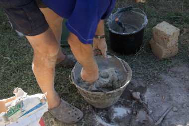 Yaşlı bir tuğlacı elinde malayla bir kovaya beton yoğuruyor. İnşaat işleri ve alçak bir duvar inşaatı. İnşaat alanında çalışan bir adam.