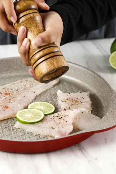 Woman Hand Grinding Pepper onto Fillet Fish on a Pan using Wooden Pepper Mill, Close up on Dori Fillet