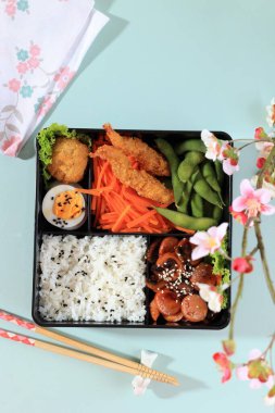 Top View Japanese Bento Lunchbox with Steamed Vegetable, Boiled Egg, Nugget, Sausage, Edamame, and Tempura. On Mint Table Background with Copy Space