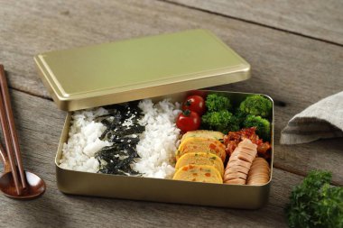 Dosirak Korean Lunch Box Contain Steamed Rice with Gyeranmari, Sausage, Kimchi, Tomato, and Steamed Broccoli. On Wooden Table