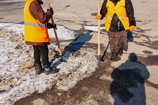 A woman worker cleans the ice and removes snow from paving slabs using an icebreaker. A man breaks ice with a steel blade crusher, an ice-breaking tool. The janitor cleans up the area.