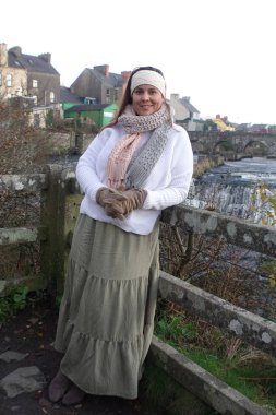 Lady wearing winter long skirt, with white jumper, knitted headband and knitted scarf to keep herself warm and spend relaxing time in nature at Ennistymon Falls in Ireland