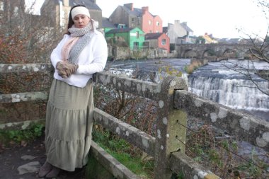 Lady wearing winter long skirt, with white jumper, knitted headband and knitted scarf to keep herself warm and spend relaxing time in nature at Ennistymon Falls in Ireland