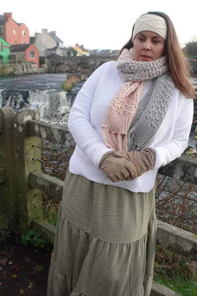 Lady wearing winter long skirt, with white jumper, knitted headband and knitted scarf to keep herself warm and spend relaxing time in nature at Ennistymon Falls in Ireland