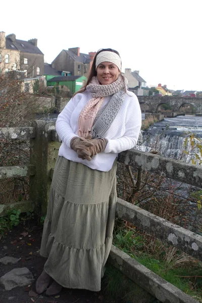 Lady wearing winter long skirt, with white jumper, knitted headband and knitted scarf to keep herself warm and spend relaxing time in nature at Ennistymon Falls in Ireland