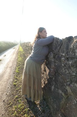 Lady leaning against a stone wall on a country road
