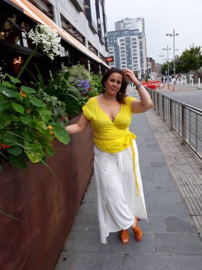 Kadın sarı yazlık bluz giyiyor, beyaz pantolon giyiyor ve çiçeklerin yanında duruyor.