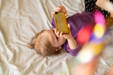 Tatlı küçük kız Z akıllı telefonda online uygulama kullanıyor, internette geziniyor, video oyunları oynuyor, selfie çekiyor, arama yapıyor, çizgi film izliyor, evdeki yatak odasında dinleniyor.