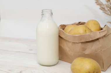 Beyaz ahşap arka planda cam bir şişede patates sütü. Yeni, taze patatesler masada kese kağıdının içinde..