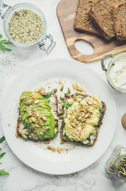 Kahvaltıda sağlıklı avokado tostları ya da öğle yemeğinde çavdar ekmeği, krem peynir, roka, dilimlenmiş avokado, balkabağı, kenevir ve susam tohumu, tuz ve biber. Vejetaryen gıda konsepti. Temiz yemek. Üst görünüm.