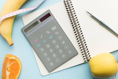 Boş kağıt defter, hesap makinesi 1200 numara, kalem, mezura ve mavi arka planda meyveler. Sağlıklı beslenme konsepti - günlük besin alımını hesapla. Üst görünüm.