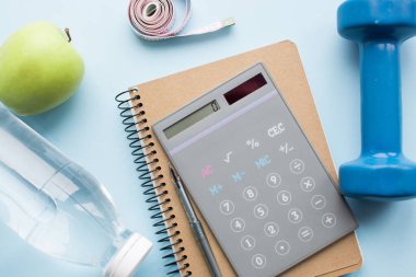 Kağıt defter, hesap makinesi, kalem, bir şişe su, Dunbbell, bant ve elma mavi arka planda. Kilo kaybı, zindelik, kalori sayımı günlük besin alımını hesaplamak. Üst görünüm.