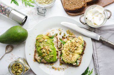 Kahvaltıda sağlıklı avokado tostları ya da öğle yemeğinde çavdar ekmeği, krem peynir, roka, dilimlenmiş avokado, balkabağı, kenevir ve susam tohumu, tuz ve biber. Vejetaryen gıda konsepti. Temiz yemek. Üst görünüm.