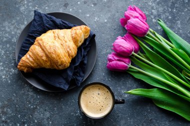 Bir fincan kahve, pembe laleler ve siyah arka planda kruvasan. Sevgililer Günü, Anneler, Kadınlar Günü sabahı konsepti. Sevgililer Günü kahvaltısı. Üst görünüm.