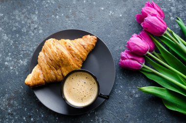Bir fincan kahve, pembe laleler ve siyah arka planda kruvasan. Sevgililer Günü, Anneler, Kadınlar Günü sabahı konsepti. Sevgililer Günü kahvaltısı. Üst görünüm.