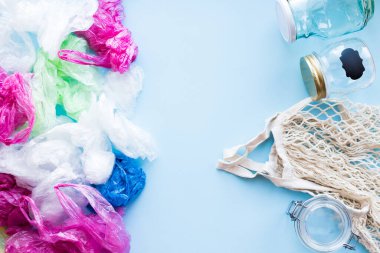 Bir sürü tek kullanımlık plastik poşet, yeniden kullanılabilir pamuk torbası ve mavi arka planda cam kavanozlara karşı. Sıfır atık. Sürdürülebilir yaşam, plastiksiz konsept. Eko dostu plastik eşyalar. Üst görünüm.