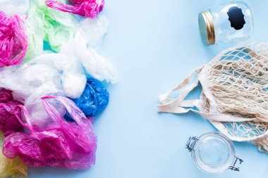 Bir sürü tek kullanımlık plastik poşet, yeniden kullanılabilir pamuk torbası ve mavi arka planda cam kavanozlara karşı. Sıfır atık. Sürdürülebilir yaşam, plastiksiz konsept. Eko dostu plastik eşyalar. Üst görünüm.
