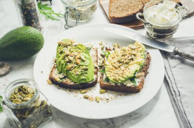 Kahvaltıda sağlıklı avokado tostları ya da öğle yemeğinde çavdar ekmeği, krem peynir, roka, dilimlenmiş avokado, balkabağı, kenevir ve susam tohumu, tuz ve biber. Vejetaryen sandviçler. Temiz yemek..