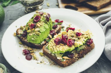 Kahvaltıda sağlıklı avokado tostları ya da öğle yemeğinde çavdar ekmeği, krem peynir, roka, dilimlenmiş avokado, kızılcık, balkabağı, kenevir ve susam tohumu. Vejetaryen sandviçler. Temiz yeme kavramı.