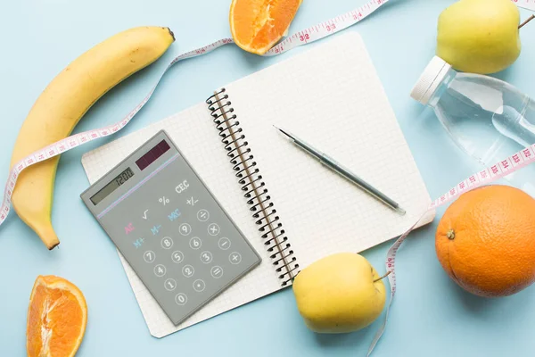 Boş kağıt defter, 1200 numaralı hesap makinesi, kalem, su şişesi, mezura ve mavi arka planda meyveler. Sağlıklı beslenme konsepti - günlük besin alımını hesapla. Üst görünüm.