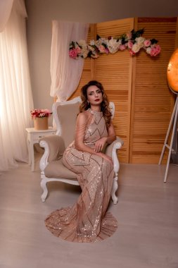 Kahverengi gözlü esmer. Pencerenin yanında. Sandalyede poz vermek. Güzel elbise. .