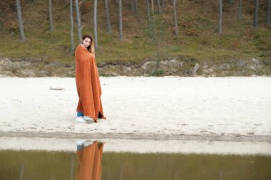 Kız deniz kıyısında dikiliyor. Arka planda bir orman var. Battaniyeli kız. Güzel kız denizin kenarında duruyor ve şafağı seyrediyor. Seyahat eden bir kız. İnanılmaz manzaralı açık hava manzarası. Deniz kıyısında güzel bir şafak. Macera yaşam tarzı