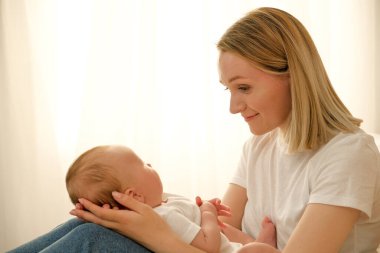 Anne kucağında yeni doğmuş bir bebek tutuyor. Arka planda güneş pencereden parlıyor..