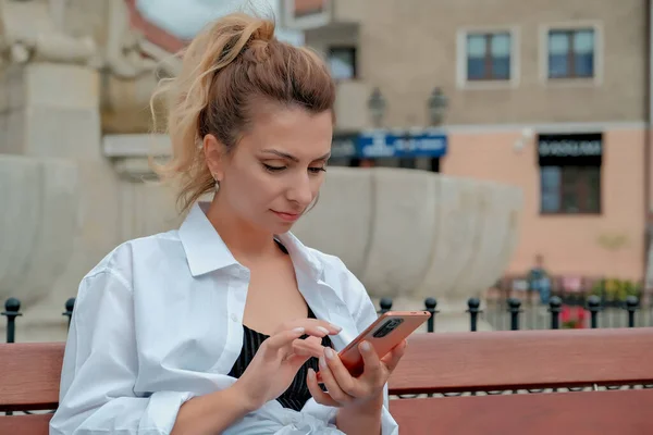 a beautiful girl sits on a bench and holds a phone in her hands. the girl orders food through the phone. the girl is talking on the phone laughing , happy , surprised .