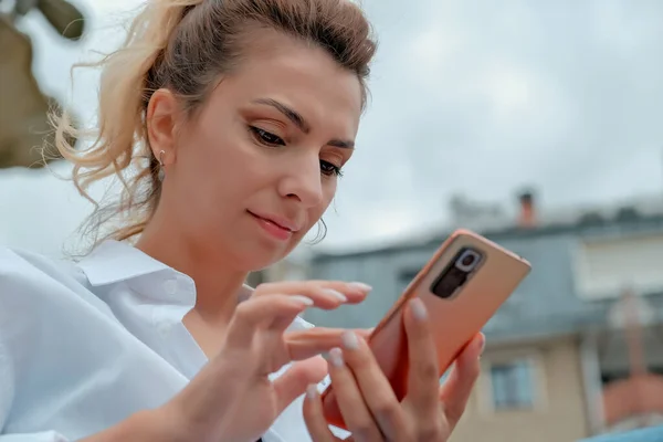 a beautiful girl sits on a bench and holds a phone in her hands. the girl orders food through the phone. the girl is talking on the phone laughing , happy , surprised .