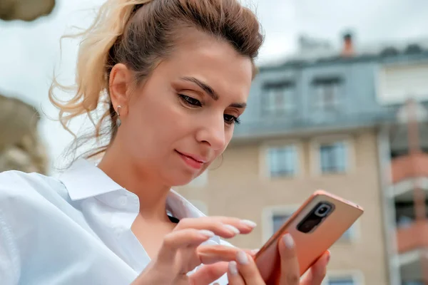 a beautiful girl sits on a bench and holds a phone in her hands. the girl orders food through the phone. the girl is talking on the phone laughing , happy , surprised .