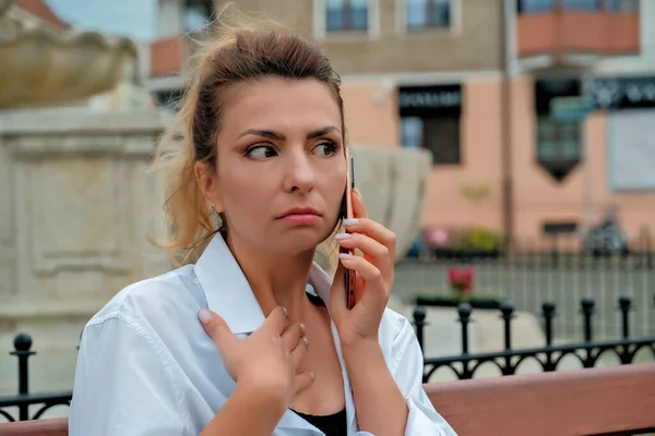 a beautiful girl sits on a bench and holds a phone in her hands. the girl orders food through the phone. the girl is talking on the phone laughing , happy , surprised .