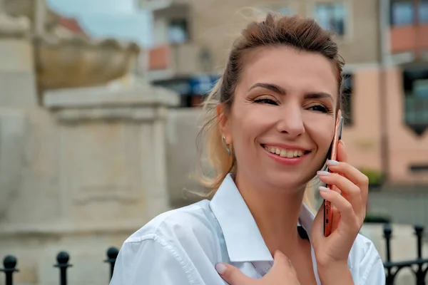 a beautiful girl sits on a bench and holds a phone in her hands. the girl orders food through the phone. the girl is talking on the phone laughing , happy , surprised .