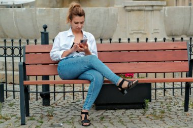 Güzel bir kız bankta oturuyor ve elinde bir telefon tutuyor. Kız telefonla yemek sipariş ediyor. Kız telefonda konuşuyor, gülüyor, mutlu, şaşırmış. .