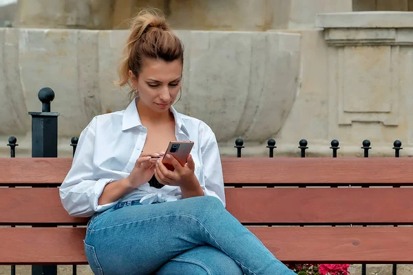 a beautiful girl sits on a bench and holds a phone in her hands. the girl orders food through the phone. the girl is talking on the phone laughing , happy , surprised .