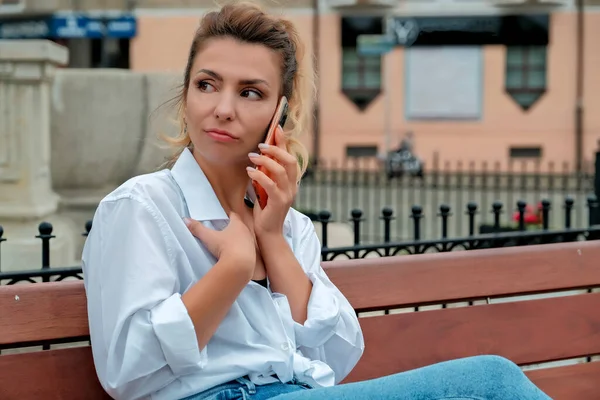 a beautiful girl sits on a bench and holds a phone in her hands. the girl orders food through the phone. the girl is talking on the phone laughing , happy , surprised .