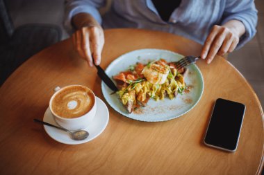 Mavi gömlekli genç kadın kahvaltıda glütensiz waffle ve cappuccino yiyor.