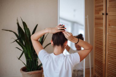 Beyaz tişörtlü, uzun saçlı esmer bir kadın evde aynanın yanında oturup kendi saçını yapıyor. Arka plan