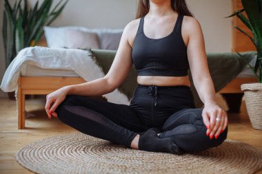 Siyah eşofmanlı genç bir kadın Lotus pozisyonunda yatağın yanındaki bir halının üzerinde oturuyor. Evde yoga yapıyor. Ev yapımı yeşil bitkiler yakınlarda duruyor. Görünmez yüz