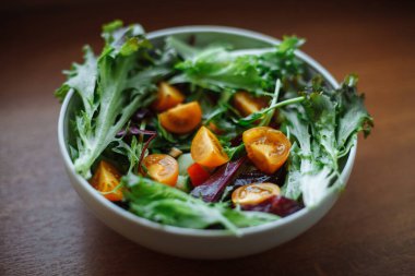 Tahta bir masada sebze salatasıyla birlikte beyaz bir tabak var. Domates, salatalık, yeşillik, marul yaprağı, biber.