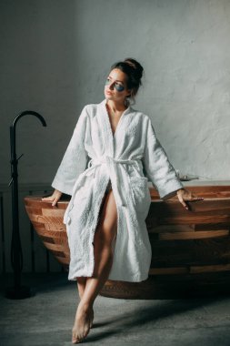 Esmer bir kadın, 30 yaşında, kafasında bir tomar para, beyaz bir bornozla banyoda, yüzünde gri lekelerle.