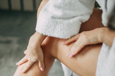 30 yaşında, uzun saçlı, beyaz bornozlu, mavi kenarlı esmer bir kadın banyoda oturuyor ve bacaklarına yağ sürüyor.