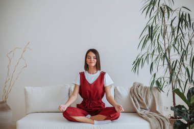 Beyaz tişörtlü, kırmızı tulumlu, esmer bir kadın beyaz bir kanepede oturma odasında yoga yapıyor. Lotus pozisyonunda oturuyor. Yakınlarda saksılarda yeşil bitkiler var.