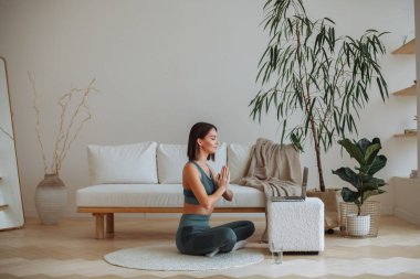 A young brunette woman in a tracksuit is doing yoga in an online lesson on a laptop at home in the living room. Sits in the lotus position with closed eyes. In the room there is a white sofa, a white rug, a white pouffe and a house plant tree in a po clipart