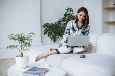 Pijamalı esmer bir kız evde beyaz bir kanepede dizüstü bilgisayar kullanıyor. Modern iç mimari