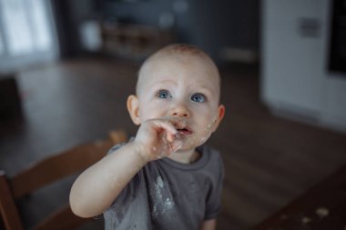 Kahverengi tişörtlü bir çocuk mutfakta bir sandalyenin üzerinde dikiliyor. Giysileri ve yüzü yiyecekle lekelenmiş.