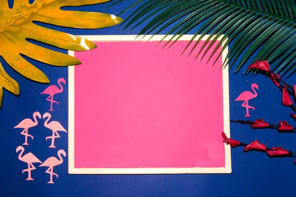 old retro frame with pink copy space on a blue background with palm leaves, flowers and pink paper flamingos, creative summer design