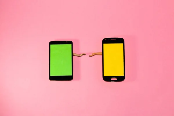 a mobile phone with a green screen offers a hand to a phone with a yellow screen that also offers a hand to him, wants to hold hands, creative idea and copy space on phones, pink background, creative